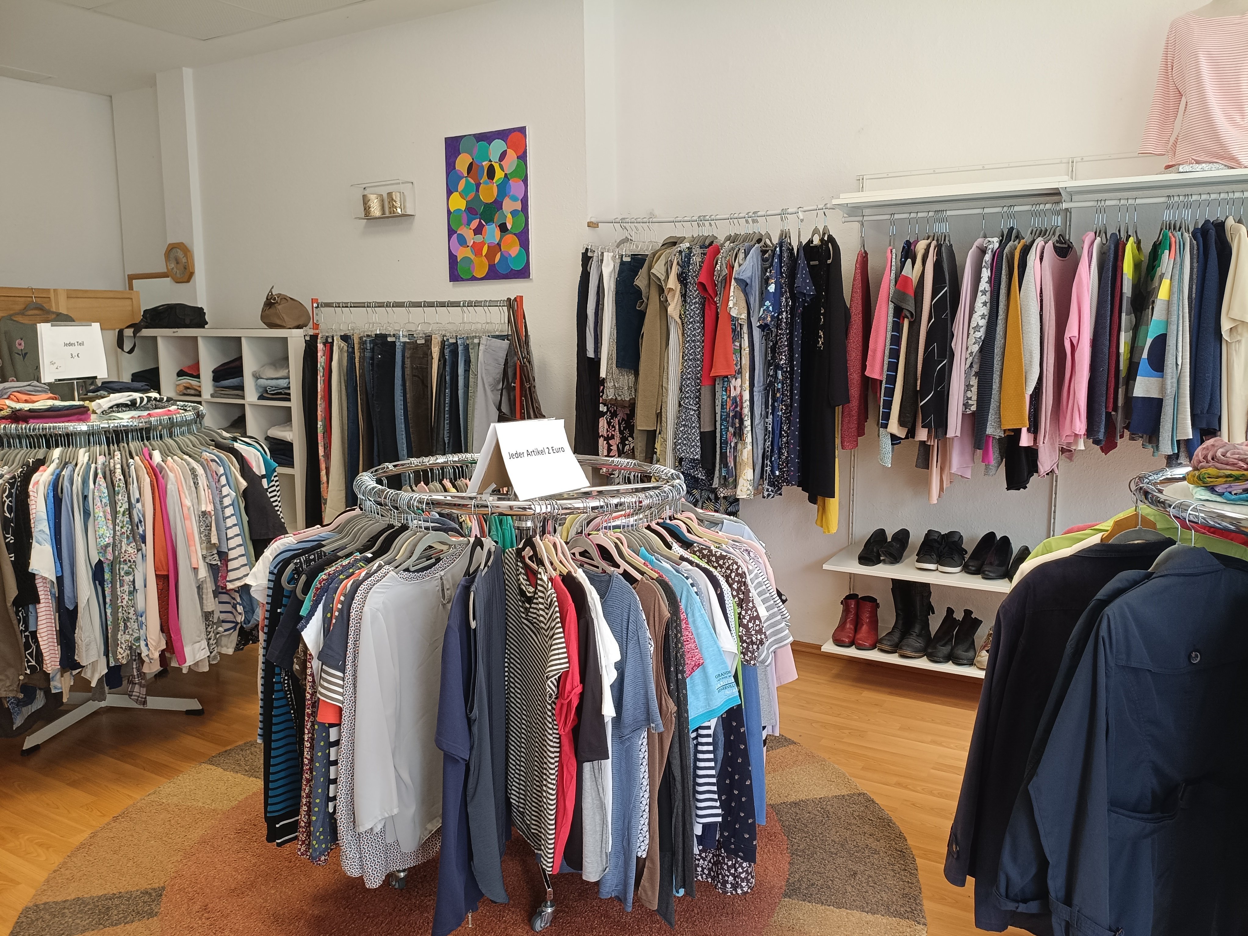 Heller Verkaufsraum des Second Design Ladens in Nordenham mit mehreren Kleiderständern voller Secondhand-Kleidung, Regalen und Schuhen. Im Vordergrund befinden sich runde Kleiderständer mit Damenbekleidung, im Hintergrund weitere Kleidungsstücke und Accessoires.