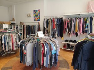 Heller Verkaufsraum des Second Design Ladens in Nordenham mit mehreren Kleiderständern voller Secondhand-Kleidung, Regalen und Schuhen. Im Vordergrund befinden sich runde Kleiderständer mit Damenbekleidung, im Hintergrund weitere Kleidungsstücke und Accessoires.