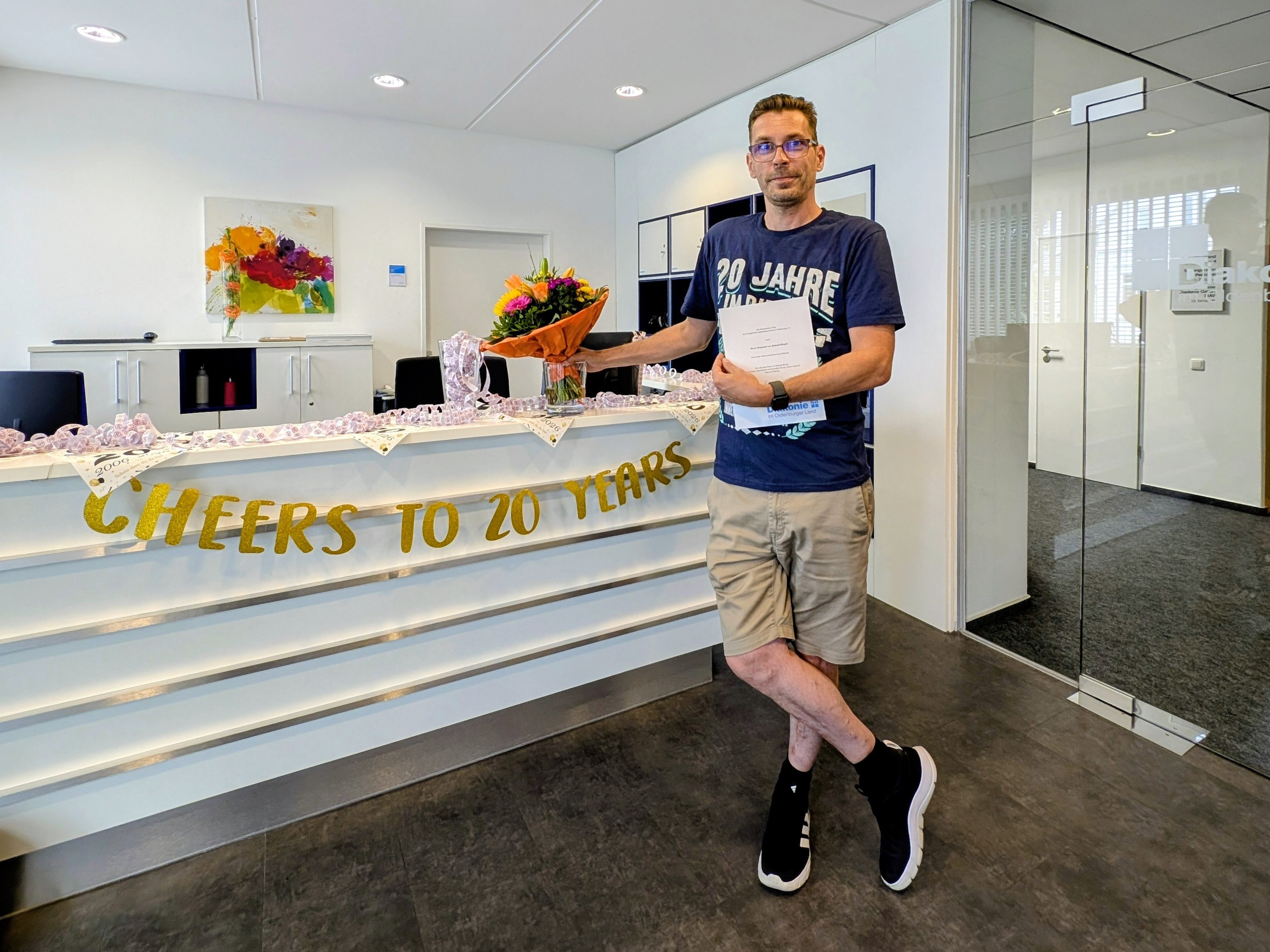 Benjamin von Bylandt steht im Eingangsbereich seines Unternehmens vor einem Empfangstresen mit der Aufschrift „Cheers to 20 Years“. Er hält einen bunten Blumenstrauß und eine Jubiläumsurkunde in den Händen. Der Empfang ist festlich dekoriert und würdigt sein 20-jähriges Dienstjubiläum.