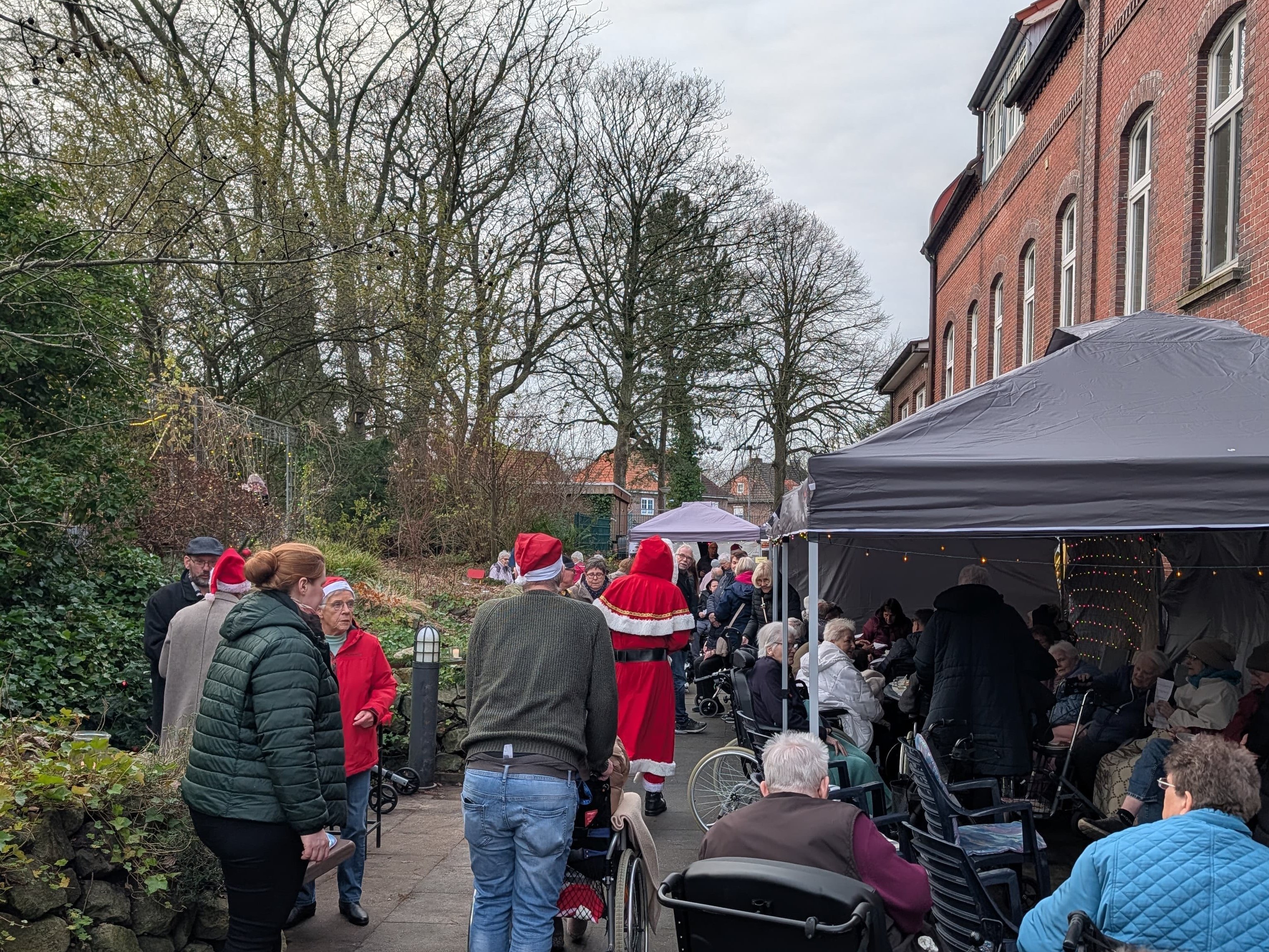Auf einem gepflasterten Weg im Freien sitzen und stehen viele ältere Menschen, einige in Rollstühlen, unter Pavillons. Eine als Weihnachtsmann verkleidete Person geht zwischen den Besucherinnen umher.