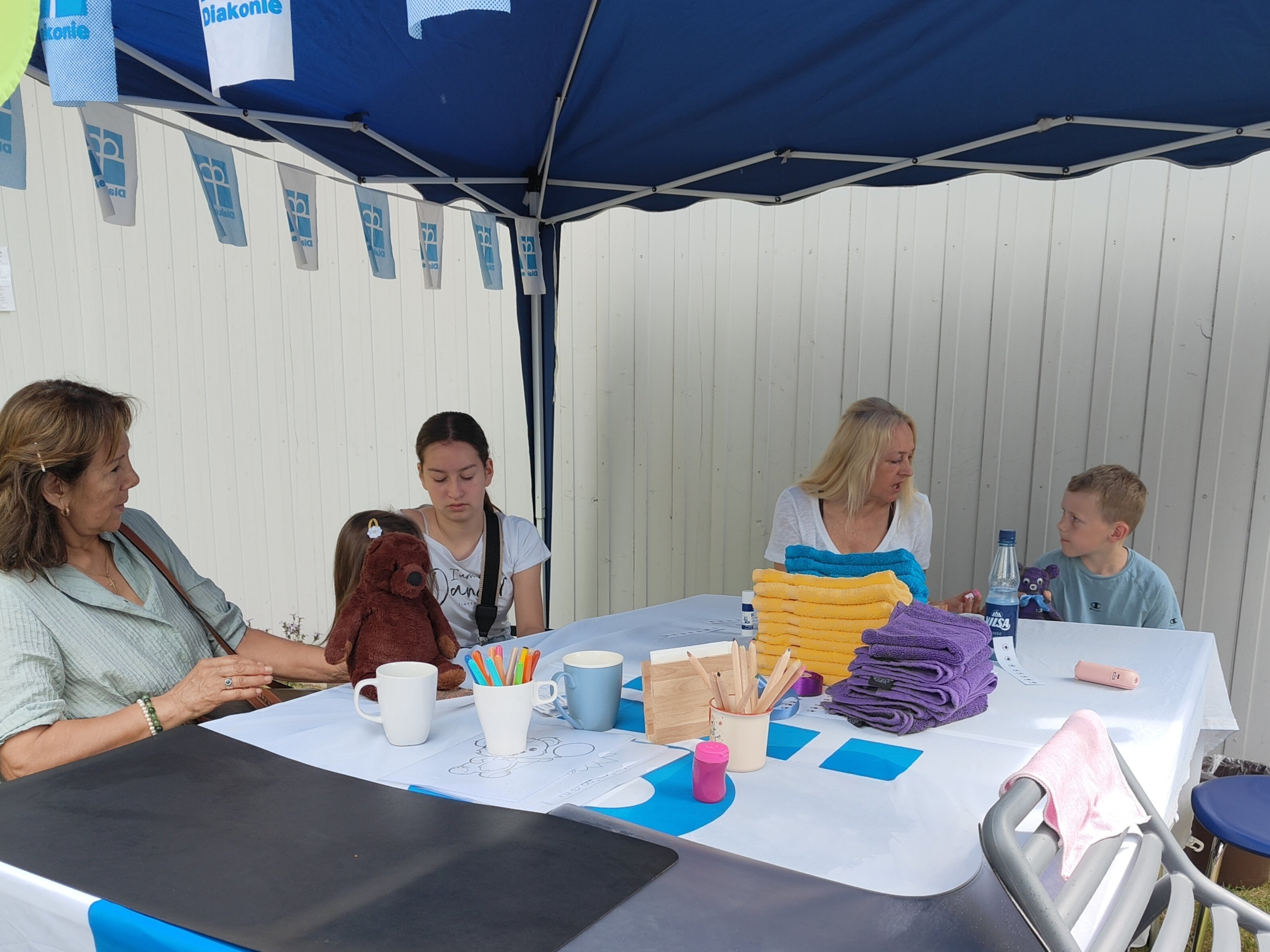 Mehrere Erwachsene und Kinder sitzen unter einem blauen Diakonie-Pavillon an einem Basteltisch. Auf dem Tisch liegen gefaltete Gästehandtücher in verschiedenen Farben sowie Bastelmaterialien. Gemeinsam werden aus den Handtüchern Teddybären gestaltet.