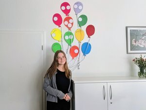 Porträt von Merle Kruse in der Jugendhilfe Collstede. Sie steht vor einer Wand mit bunten Luftballons, die die Monate des Jahres symbolisieren, und blickt lächelnd in die Kamera.