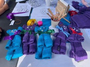 Mehrere aus bunten Gästehandtüchern gebastelte Teddybären mit Schleifen und Wackelaugen liegen auf einem Basteltisch. Im Hintergrund sind Stifte, Ausmalbilder und weiteres Bastelmaterial zu sehen.