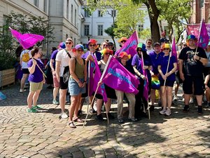 Eine große Gruppe von Menschen steht bei sonnigem Wetter auf einem Platz vor einer Backsteinkirche in Oldenburg zusammen. Viele tragen bunte Regenbogenhüte und halten lilafarbene Fahnen der Diakonie sowie Regenbogenflaggen in den Händen. Im Hintergrund ist ein Banner mit der Aufschrift „Gottes Welt ist bunt!“ zu sehen. Die Gruppe zeigt beim Christopher Street Day gemeinsam ein Zeichen für Vielfalt und Zusammenhalt.