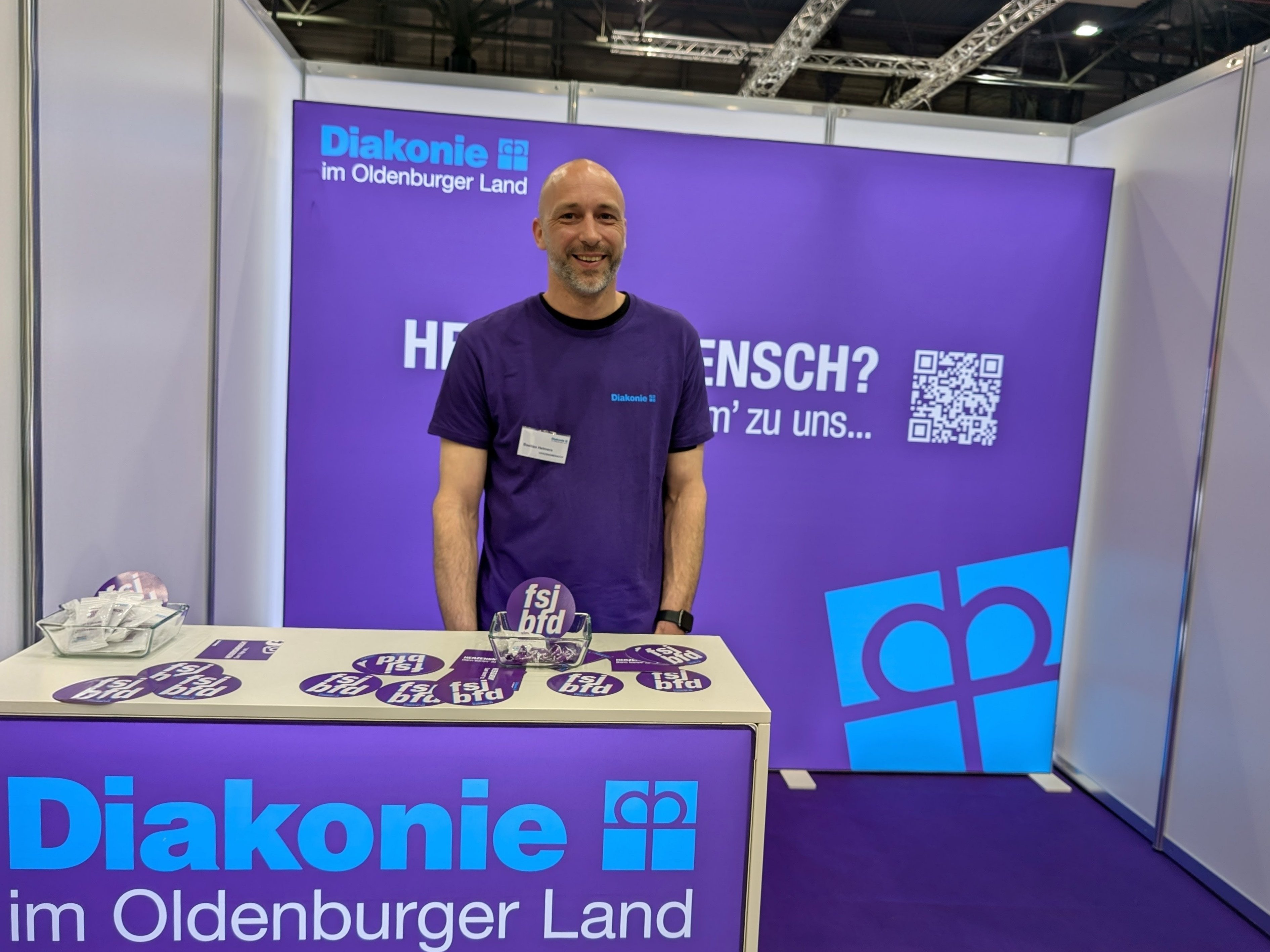 Mitarbeiter der Diakonie im Oldenburger Land steht an einem Messestand in den Weser-Ems-Hallen in Oldenburg. Der Stand ist in Lila und Blau gestaltet und informiert über Ausbildungs-, Studien- und Berufsmöglichkeiten sowie Freiwilligendienste bei der Diakonie.