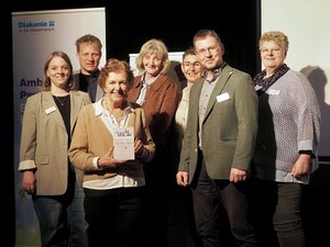 Gruppenfoto nach einer Lesung im Theater Fatale: Die Autorin Helga Winkler steht in der Mitte und hält ihr Buch „Bye bye, Balu“ in den Händen. Um sie herum stehen Vertreter*innen der Diakonie, des Fördervereins „Stationäres Hospiz Wesermarsch e.V.“ sowie der ambulanten Hospizdienste Nordenham und umzu. Die Gruppe blickt in die Kamera und lächelt. Im Hintergrund ist ein Banner der Diakonie zu sehen.