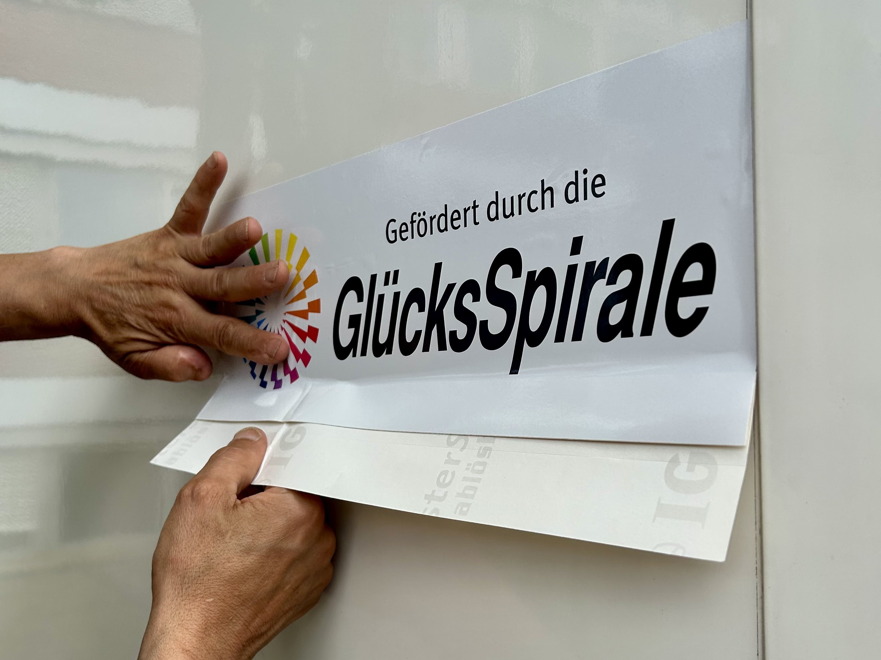 Zwei Hände bringen den Aufkleber „Gefördert durch die GlücksSpirale“ an der Seitenwand des neuen VW Crafter des Möbeldienstes der Diakonie im Oldenburger Land an.
