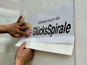 Zwei Hände bringen den Aufkleber „Gefördert durch die GlücksSpirale“ an der Seitenwand des neuen VW Crafter des Möbeldienstes der Diakonie im Oldenburger Land an.