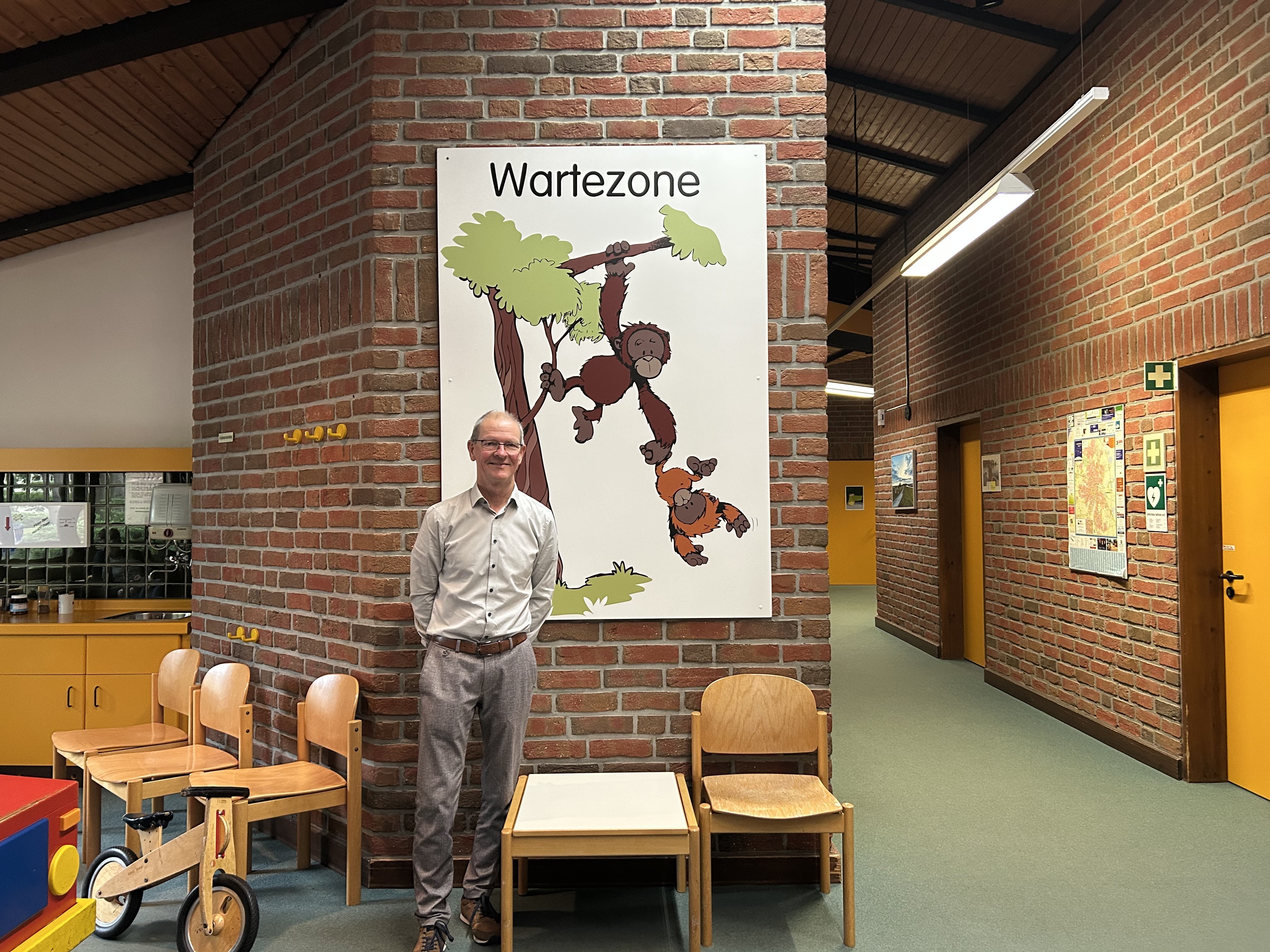 Dr. Michael Wagner steht lächelnd im Wartebereich des Sozialpädiatrischen Zentrums vor einem Wandbild mit zwei Orang-Utans und dem Schriftzug „Wartezone“.