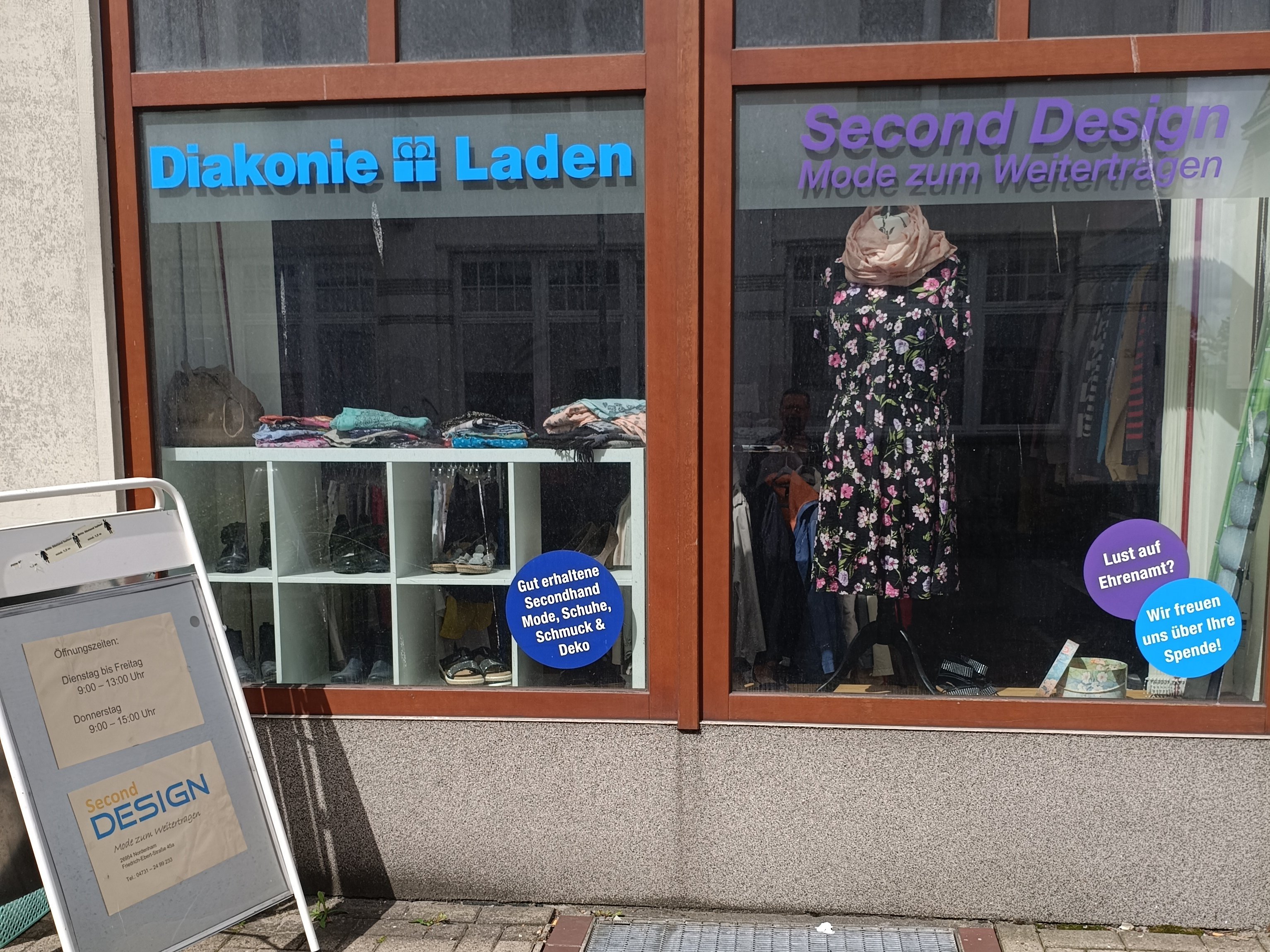 Schaufensterfront des Second Design Ladens in Nordenham mit den Schriftzügen „Diakonie Laden“ und „Second Design – Mode zum Weitertragen“. Im Schaufenster sind Secondhand-Kleidung, Schuhe und Dekoration ausgestellt, vor dem Geschäft steht ein Aufsteller mit den Öffnungszeiten.