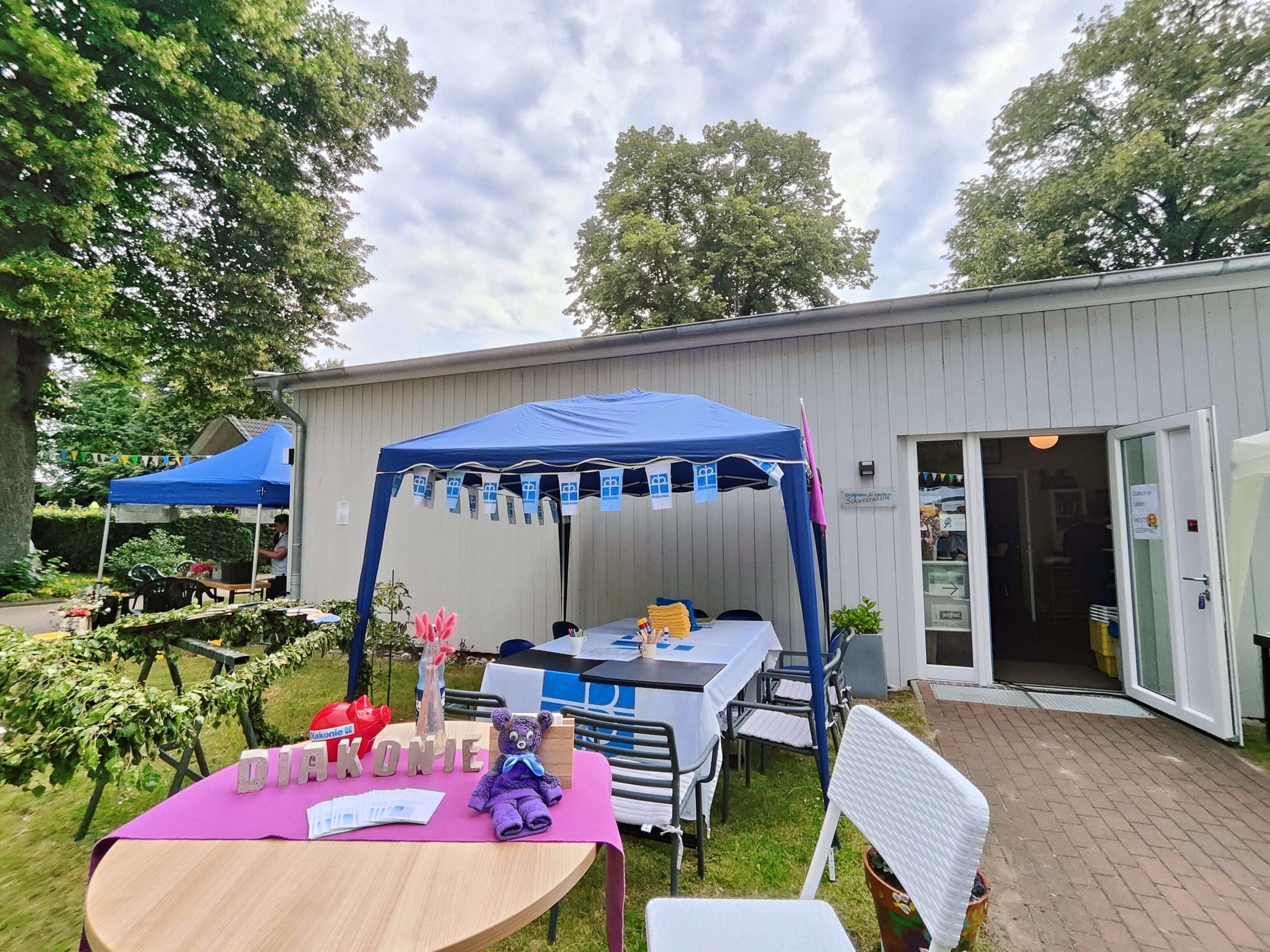 Ein blauer Pavillon der Diakonie mit Tischen und Bastelmaterial steht vor einem Gebäude mit geöffnetem Eingang zum Diakonieladen. Im Vordergrund sind ein dekorierter Tisch mit Informationsmaterial und ein aus Gästehandtüchern gebastelter Teddybär zu sehen.