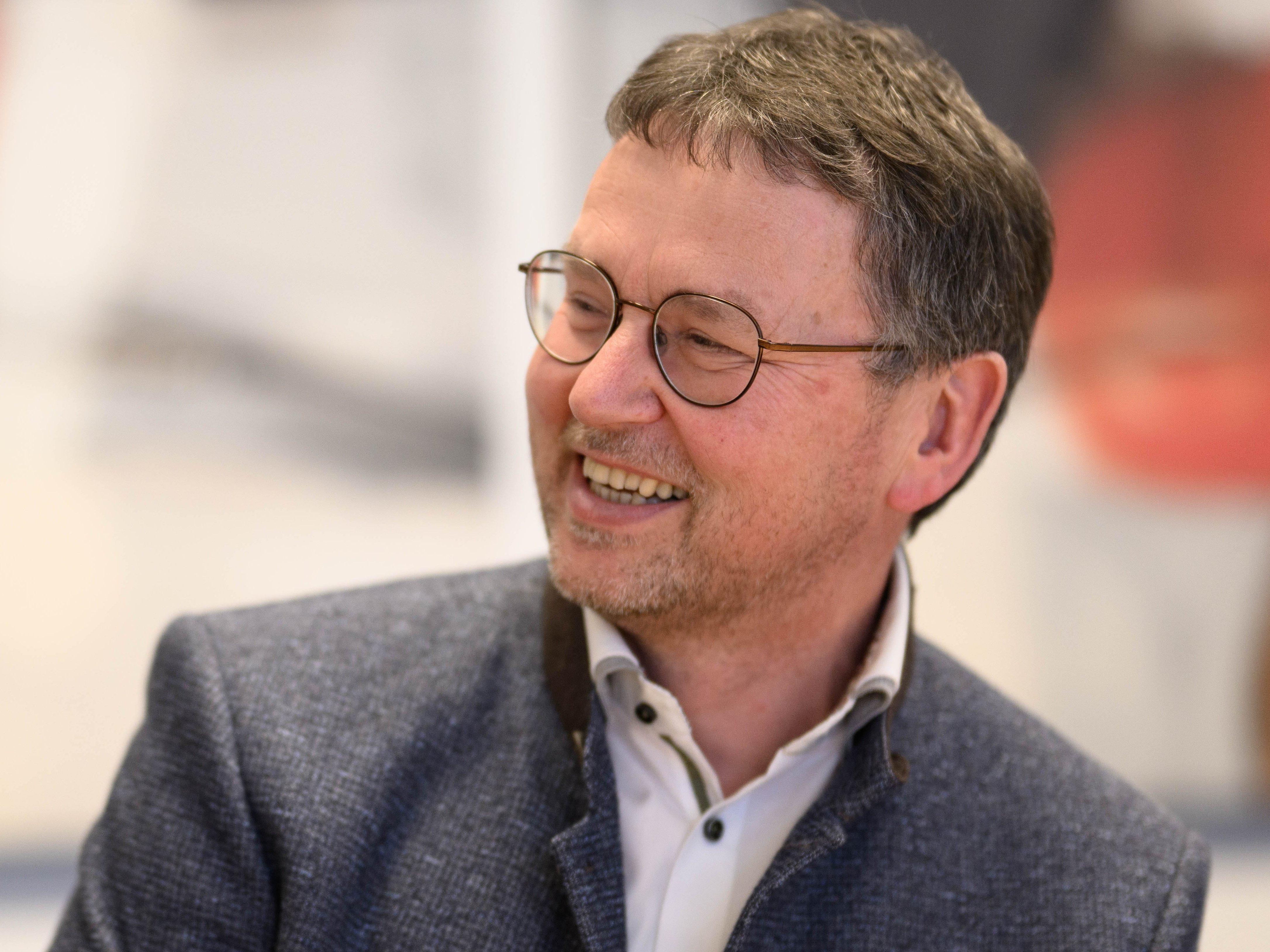 Thomas Scheurenbrand lächelt und blickt zur Seite. Er trägt eine runde Brille, ein weißes Hemd und ein dunkelblaues Sakko mit Namensschild. Der Hintergrund ist unscharf und zeigt einen hellen Innenraum.