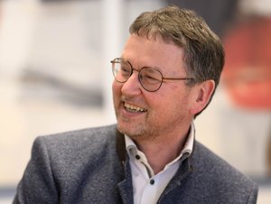 Thomas Scheurenbrand lächelt und blickt zur Seite. Er trägt eine runde Brille, ein weißes Hemd und ein dunkelblaues Sakko mit Namensschild. Der Hintergrund ist unscharf und zeigt einen hellen Innenraum.
