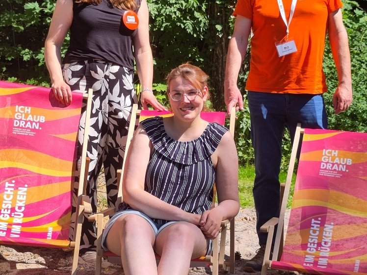 Denise Irmscher, Maike Hamacher und Marius Blümel präsentieren die Aktion von Brot für die Welt auf der Landesgartenschau in Bad Nenndorf. Sie stehen und sitzen zwischen drei Liegestühlen mit der Aufschrift „Ich glaub dran“ im Freien.