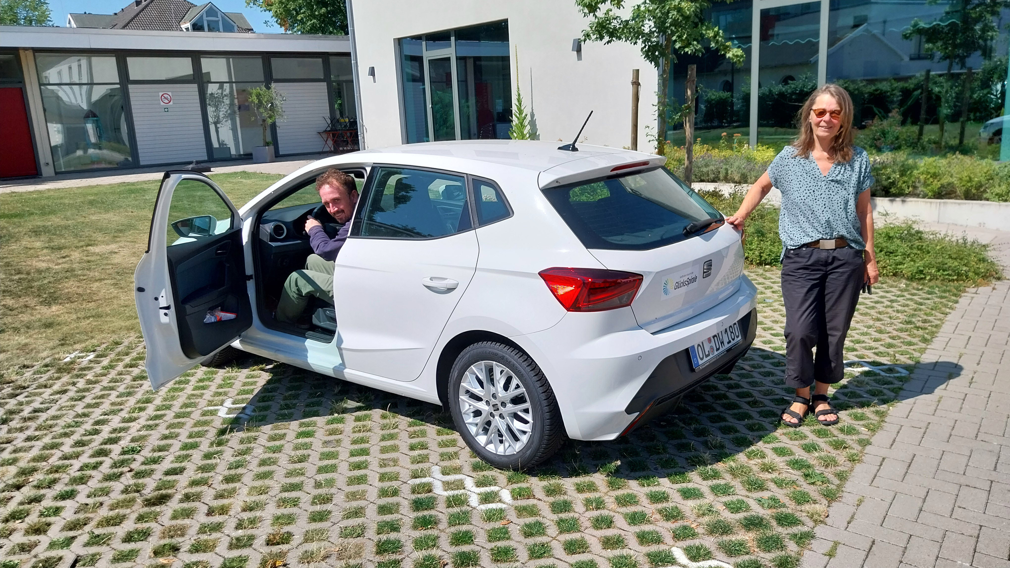 Zwei Mitarbeitende der Straffälligenhilfe der Diakonie im Oldenburger Land stehen an einem weißen Seat Ibiza mit Förderhinweis der GlücksSpirale vor einem Dienstgebäude.