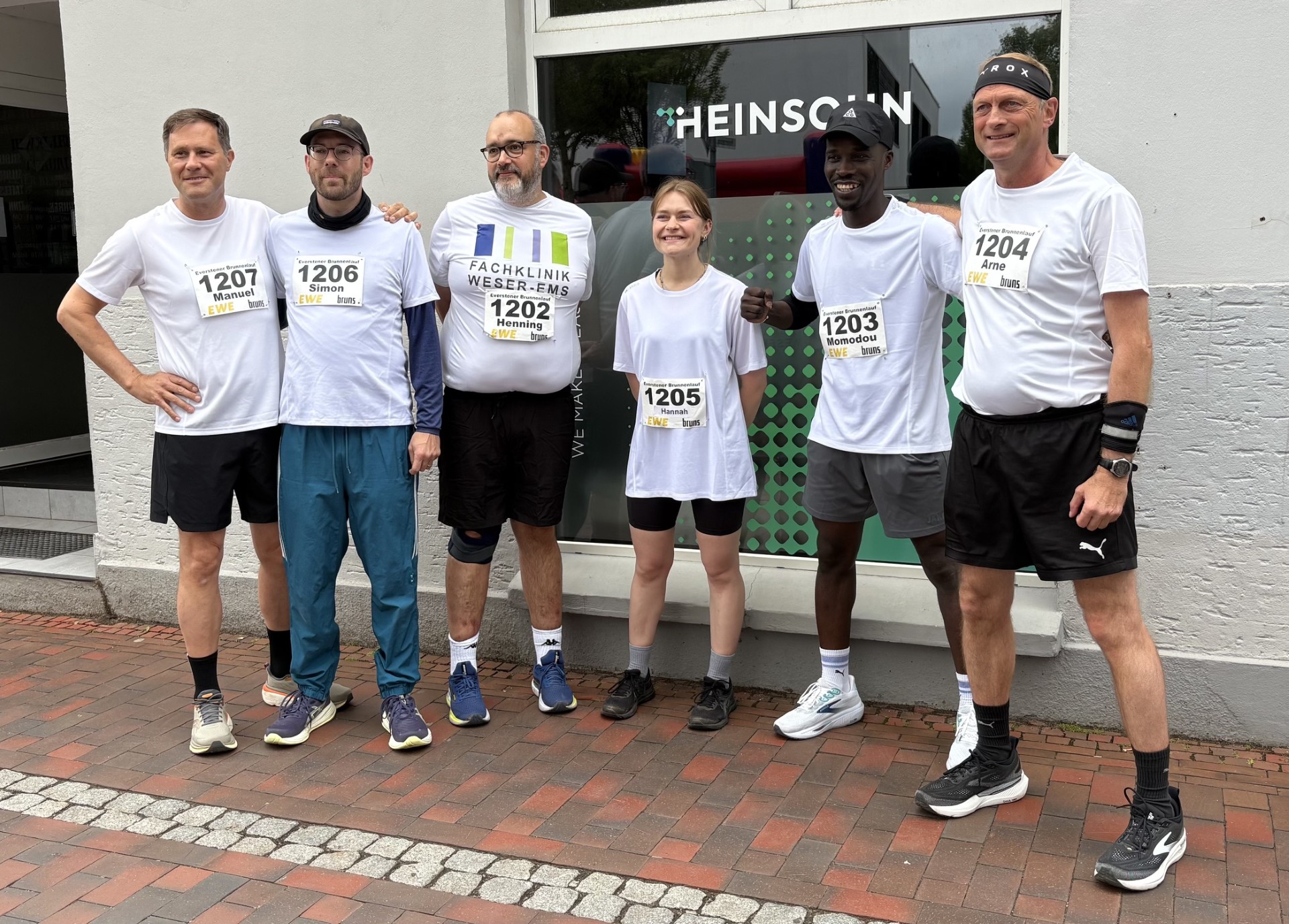 Sechs Mitglieder des Lauf-Teams der Fachklinik Weser-Ems posieren vor ihrer Teilnahme am Everstener Brunnenlauf für ein Gruppenfoto. Sie tragen weiße Laufshirts mit Startnummern und wirken motiviert und gut gelaunt.