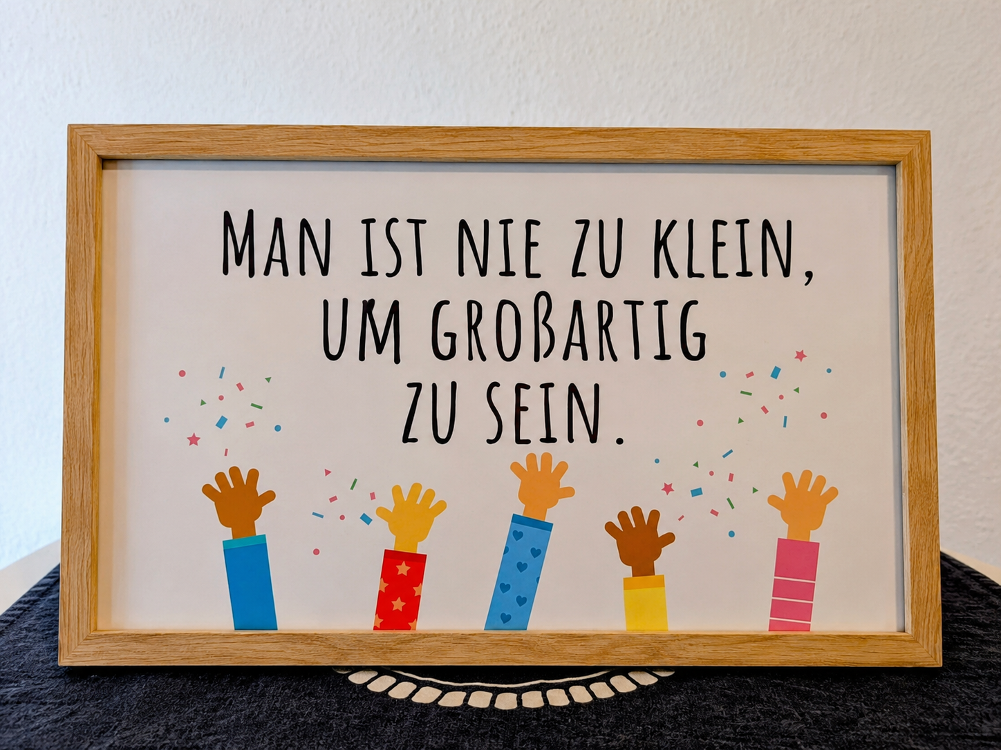 Gerahmtes Bild mit dem Spruch „Man ist nie zu klein, um großartig zu sein“ und bunten Kinderhänden.