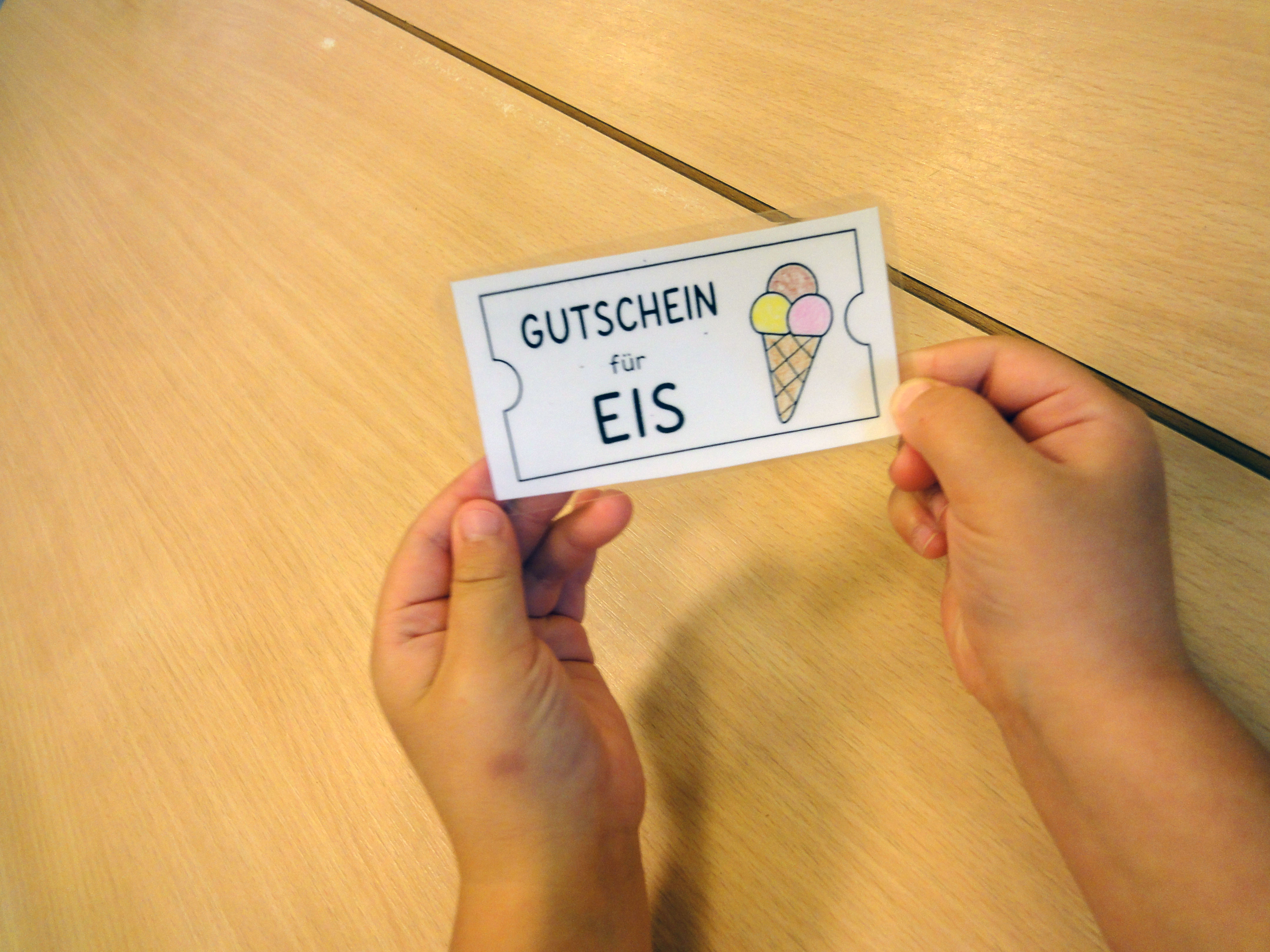 Ein Kind hält einen selbst gestalteten Eisgutschein mit einer bunten Eiswaffel in den Händen. Der Gutschein wurde für eine Eisaktion der Elefantengruppe in der DiKiTa zum Start der Sommerferien gestaltet.